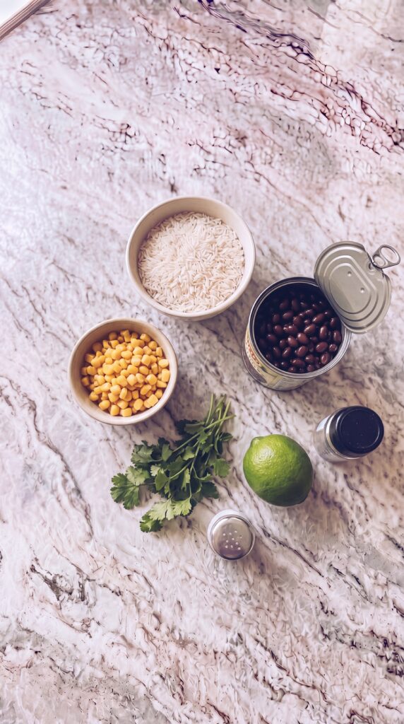 Easy High Protein Burrito Bowls For Meal Prep burrito bowl ingredients including canned black beans rice and corn arranged on marble countertop